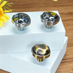 May include: Three cabinet knobs with decorative designs. Two knobs have a shiny, silver finish with a textured, irregular top and a silver base. The third knob has a bronze finish with a similar textured top and a bronze base. The knobs are displayed on a white surface.