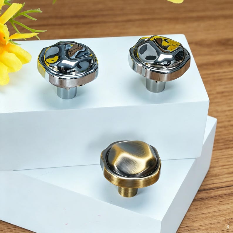 May include: Three cabinet knobs with decorative designs. Two knobs have a shiny, silver finish with a textured, irregular top and a silver base. The third knob has a bronze finish with a similar textured top and a bronze base. The knobs are displayed on a white surface.