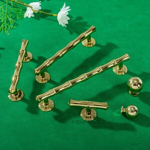 May include: A selection of gold-coloured bamboo-style cabinet hardware, including handles and knobs, arranged on a green surface. The hardware has a shiny finish and is designed to resemble bamboo stalks. The image also includes white flowers.