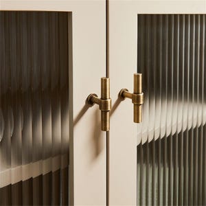 K&ouml;nnte beinhalten: Nahaufnahme einer Schrankt&uuml;r mit geriffelten Glasscheiben und antiker messingfarbener Hardware. Die Schrankt&uuml;ren sind cremefarben und die Griffe sind T-f&ouml;rmig.