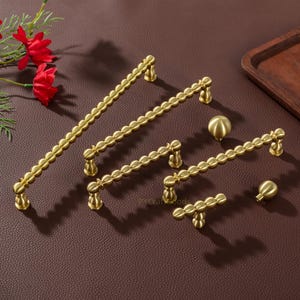 K&ouml;nnte beinhalten: Eine Auswahl an goldfarbenen Schrankbeschl&auml;gen, darunter Griffe und Kn&ouml;pfe in verschiedenen Gr&ouml;&szlig;en. Die Griffe haben ein wellenf&ouml;rmiges Design, und die Kn&ouml;pfe sind kugelf&ouml;rmig. Auf einer braunen Oberfl&auml;che angeordnet, mit roten Blumen im Hintergrund.