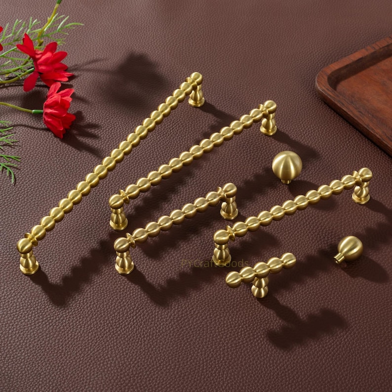 K&ouml;nnte beinhalten: Eine Auswahl an goldfarbenen Schrankbeschl&auml;gen, darunter Griffe und Kn&ouml;pfe in verschiedenen Gr&ouml;&szlig;en. Die Griffe haben ein wellenf&ouml;rmiges Design, und die Kn&ouml;pfe sind kugelf&ouml;rmig. Auf einer braunen Oberfl&auml;che angeordnet, mit roten Blumen im Hintergrund.