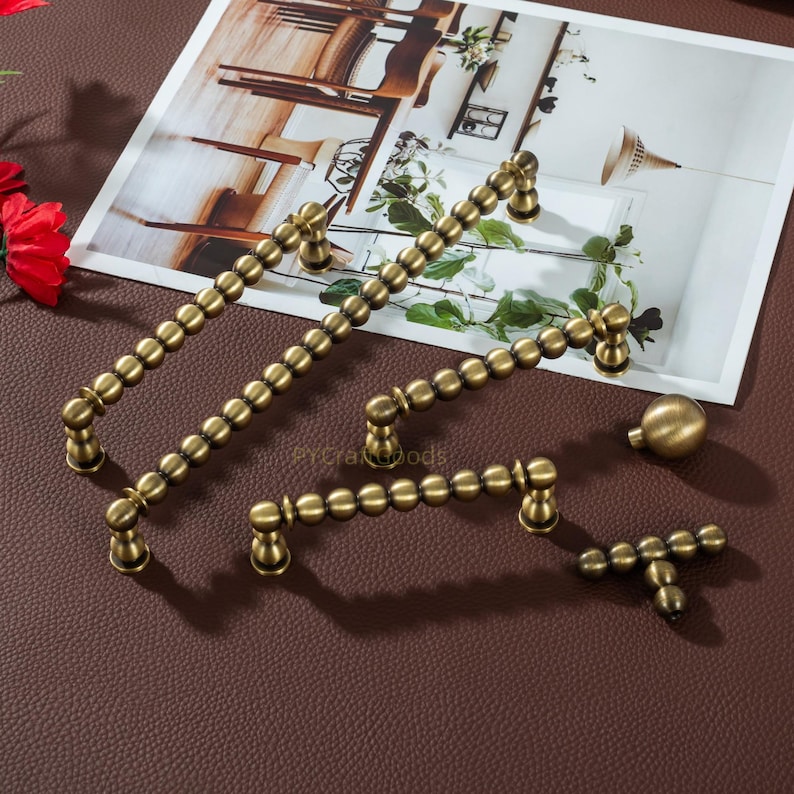 K&ouml;nnte beinhalten: Antikes Messing-Schrankbeschlagsset. Das Set enth&auml;lt verschiedene Gr&ouml;&szlig;en von Griffen und einen Knauf. Die Griffe bestehen aus kleinen, verbundenen Kugeln. Die Hardware wird auf einer braunen Oberfl&auml;che mit einem Foto im Hintergrund pr&auml;sentiert.