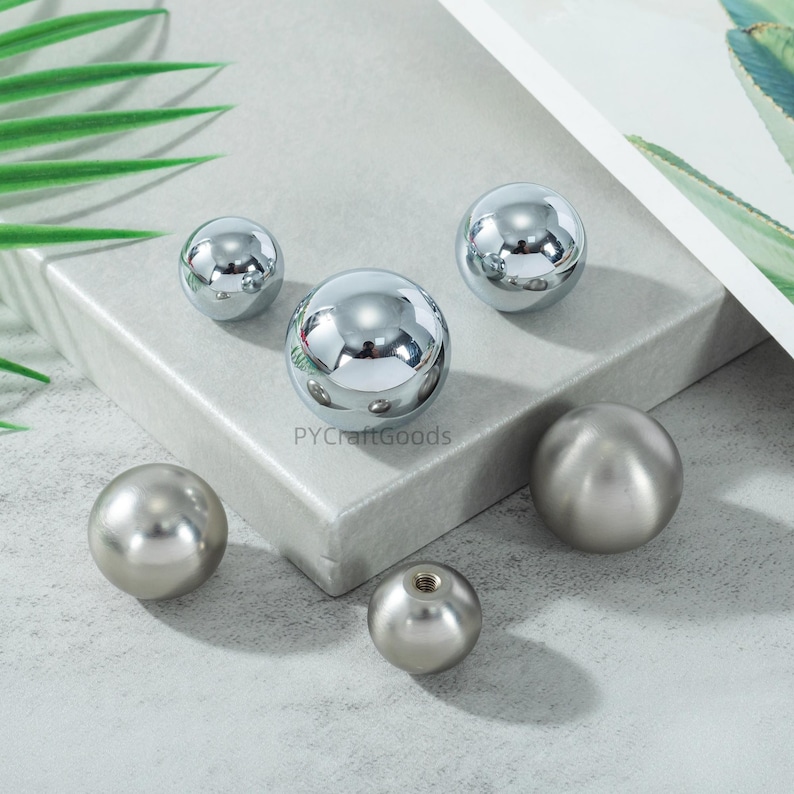 K&ouml;nnte beinhalten: Eine Sammlung von sechs Metallkugeln in verschiedenen Gr&ouml;&szlig;en. Einige Kugeln haben eine gl&auml;nzende, reflektierende Chromoberfl&auml;che, andere ein mattes, geb&uuml;rstetes Metallaussehen. Die Kugeln sind auf einer hellgrauen Oberfl&auml;che angeordnet. Ma&szlig;e in Zentimetern.