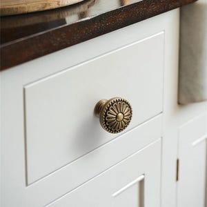 K&ouml;nnte beinhalten: Nahaufnahme eines wei&szlig;en Schranks mit einem dekorativen Messingknauf. Der Knauf hat ein Blumenmuster und ist an einer wei&szlig;en Schublade befestigt. Der Schrank ist Teil einer K&uuml;che oder eines Badezimmers.