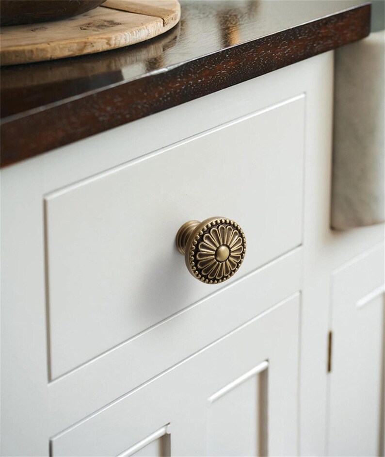 K&ouml;nnte beinhalten: Nahaufnahme eines wei&szlig;en Schranks mit einem dekorativen Messingknauf. Der Knauf hat ein Blumenmuster und ist an einer wei&szlig;en Schublade befestigt. Der Schrank ist Teil einer K&uuml;che oder eines Badezimmers.