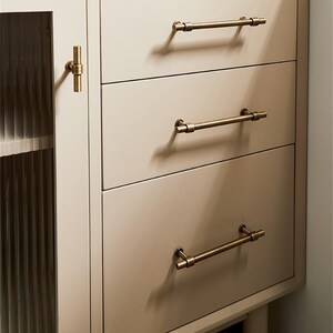 K&ouml;nnte beinhalten: Ein cremefarbener Schrank mit drei Schubladen und einer Glast&uuml;r. Die Schubladen haben horizontale, antik-messingfarbene Stangengriffe. Die Schrankt&uuml;r hat einen passenden messingfarbenen Knopf. Der Schrank ist neutralfarben und hat ein modernes Design.