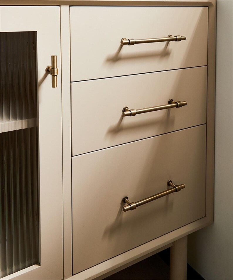 K&ouml;nnte beinhalten: Ein cremefarbener Schrank mit drei Schubladen und einer Glast&uuml;r. Die Schubladen haben horizontale, antik-messingfarbene Stangengriffe. Die Schrankt&uuml;r hat einen passenden messingfarbenen Knopf. Der Schrank ist neutralfarben und hat ein modernes Design.