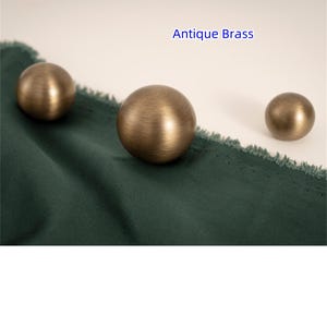 K&ouml;nnte beinhalten: Drei antike Messingkugeln unterschiedlicher Gr&ouml;&szlig;e liegen auf einem dunkelgr&uuml;nen Stoff. Die Kugeln haben eine warme, metallische Oberfl&auml;che. Der Text "Antique Brass" ist oben im Bild blau dargestellt.