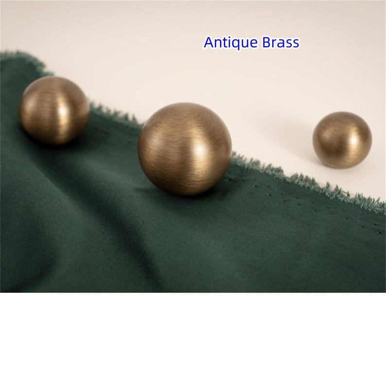 K&ouml;nnte beinhalten: Drei antike Messingkugeln unterschiedlicher Gr&ouml;&szlig;e liegen auf einem dunkelgr&uuml;nen Stoff. Die Kugeln haben eine warme, metallische Oberfl&auml;che. Der Text "Antique Brass" ist oben im Bild blau dargestellt.