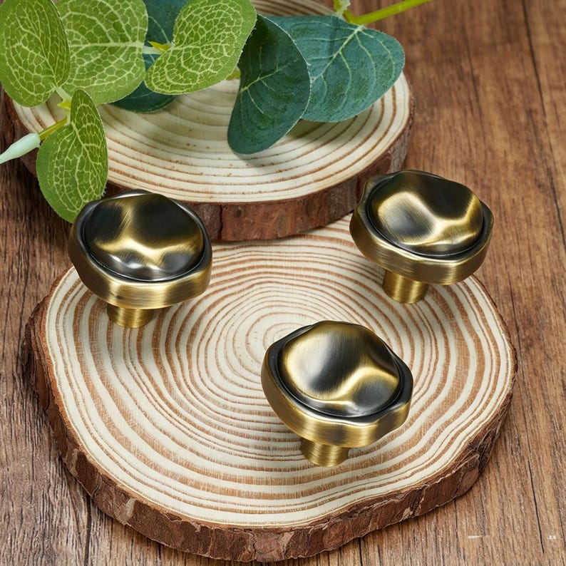 May include: Three antique brass cabinet knobs with an irregular shape. The knobs have a dark, aged finish and are mounted on a wooden surface. The knobs are likely for furniture or decorative use.