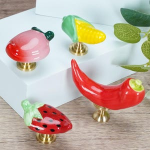 Pode incluir: Uma cole&ccedil;&atilde;o de puxadores de arm&aacute;rio de cer&acirc;mica em forma de frutas e vegetais. O conjunto inclui um p&ecirc;ssego, milho, pimenta vermelha e um morango. Cada puxador tem uma base de lat&atilde;o e &eacute; pintado &agrave; m&atilde;o com cores vibrantes.