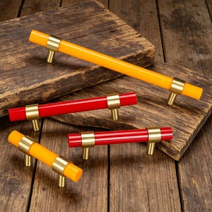 May include: A collection of cabinet pulls in vibrant colors. The pulls feature a cylindrical design with brass-colored accents. The color options include orange and red, offering a modern touch to furniture. The pulls are displayed on a wooden surface.