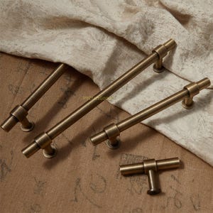 K&ouml;nnte beinhalten: Vier antike Messing-Schrankgriffe in verschiedenen Gr&ouml;&szlig;en. Die T-Stangen-Griffe sind auf runden Sockeln montiert. Die Hardware wird auf einem neutralfarbenen Stoffhintergrund pr&auml;sentiert. Die Griffe haben eine Vintage- und Industrie&auml;sthetik.