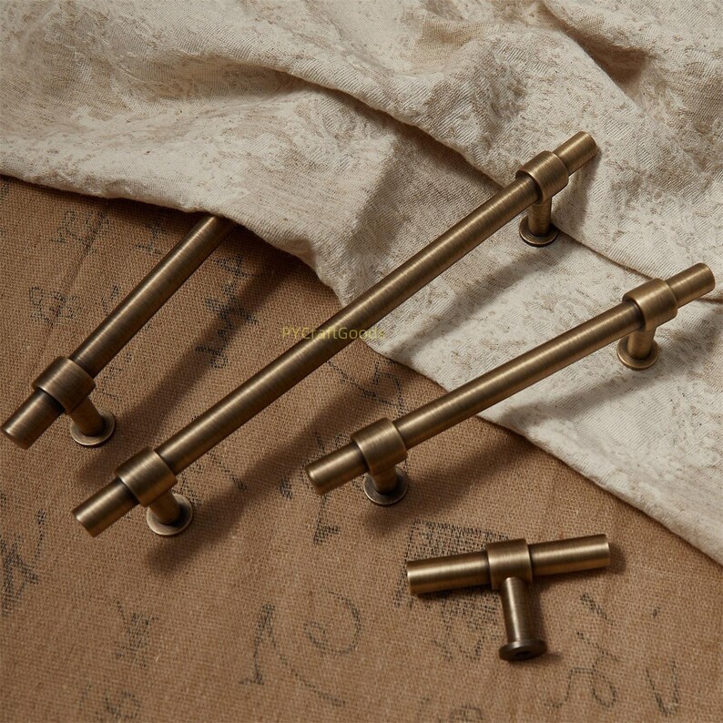 K&ouml;nnte beinhalten: Vier antike Messing-Schrankgriffe in verschiedenen Gr&ouml;&szlig;en. Die T-Stangen-Griffe sind auf runden Sockeln montiert. Die Hardware wird auf einem neutralfarbenen Stoffhintergrund pr&auml;sentiert. Die Griffe haben eine Vintage- und Industrie&auml;sthetik.