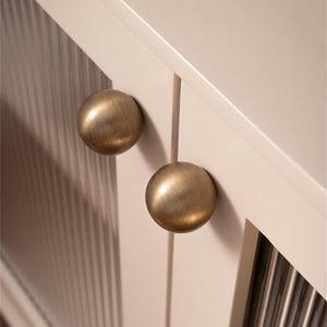 K&ouml;nnte beinhalten: Nahaufnahme einer Schrankt&uuml;r mit zwei runden Kn&ouml;pfen aus antikem Messing. Die T&uuml;r ist in einem hellen Cremeton gehalten und hat vertikale geriffelte Glaspaneele. Die Kn&ouml;pfe sind mittig angeordnet und verleihen einen klassischen, eleganten Look.