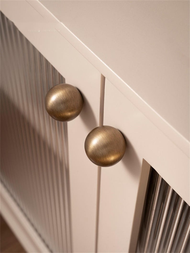 K&ouml;nnte beinhalten: Nahaufnahme einer Schrankt&uuml;r mit zwei runden Kn&ouml;pfen aus antikem Messing. Die T&uuml;r ist in einem hellen Cremeton gehalten und hat vertikale geriffelte Glaspaneele. Die Kn&ouml;pfe sind mittig angeordnet und verleihen einen klassischen, eleganten Look.