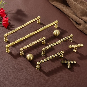 May include: A collection of brass-colored cabinet hardware, including various sizes of handles and knobs. The handles feature a beaded design, and the knobs are spherical. The hardware is displayed on a brown surface.
