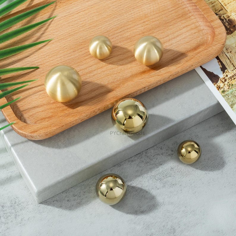 K&ouml;nnte beinhalten: Eine Sammlung goldfarbener, kugelf&ouml;rmiger Schrankkn&ouml;pfe und -griffe. Einige haben ein gerilltes Design, andere sind glatt und reflektierend. Sie sind auf einem Holztablett und einer grauen Oberfl&auml;che angeordnet.