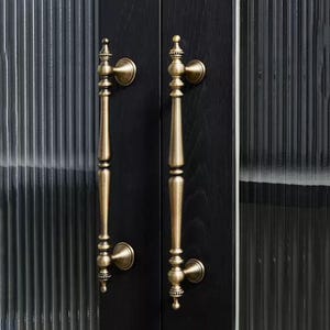 Peut inclure: Deux poign&eacute;es de porte en laiton antique avec des d&eacute;tails orn&eacute;s sont mont&eacute;es sur une porte en bois noir. Les poign&eacute;es ont un design vertical et allong&eacute; avec des fleurettes d&eacute;coratives et des plaques arri&egrave;re circulaires. La porte a un panneau de verre nervur&eacute;.