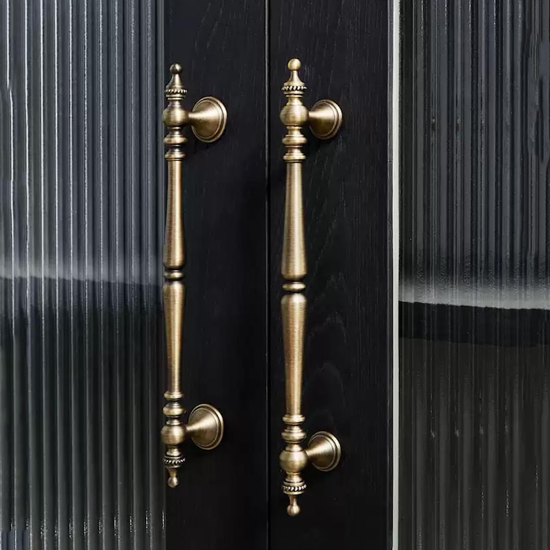 Peut inclure: Deux poign&eacute;es de porte en laiton antique avec des d&eacute;tails orn&eacute;s sont mont&eacute;es sur une porte en bois noir. Les poign&eacute;es ont un design vertical et allong&eacute; avec des fleurettes d&eacute;coratives et des plaques arri&egrave;re circulaires. La porte a un panneau de verre nervur&eacute;.