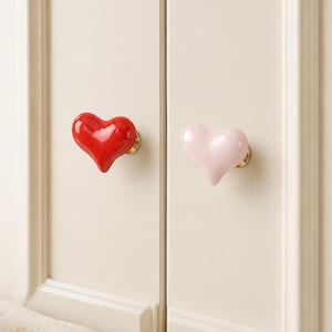 Pode incluir: Dois puxadores de arm&aacute;rio em forma de cora&ccedil;&atilde;o, um vermelho brilhante e outro rosa p&aacute;lido. Ambos est&atilde;o montados numa porta de arm&aacute;rio creme com ferragens douradas. Os puxadores adicionam um toque decorativo aos m&oacute;veis.