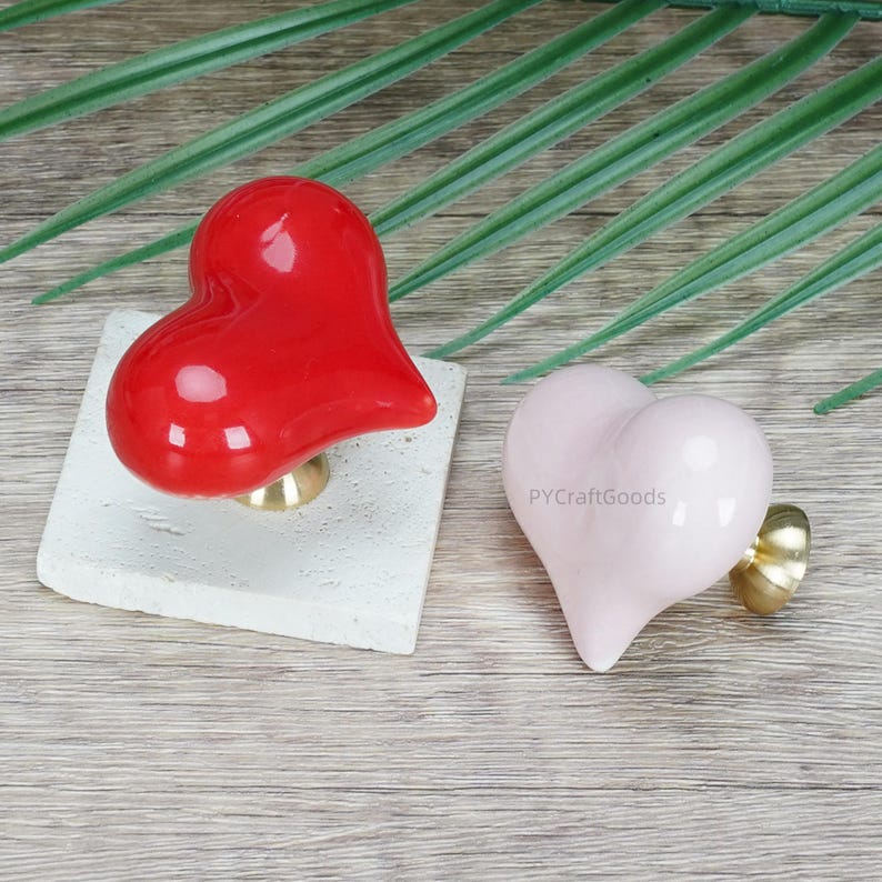 Pode incluir: Dois puxadores de arm&aacute;rio em forma de cora&ccedil;&atilde;o. Um &eacute; vermelho brilhante, montado numa base quadrada esbranqui&ccedil;ada. O outro &eacute; rosa p&aacute;lido, com uma base dourada. Ferragens decorativas para m&oacute;veis.