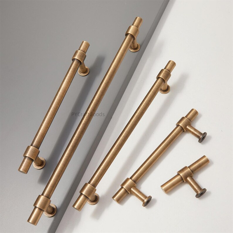 K&ouml;nnte beinhalten: Eine Auswahl an antiken Messing-Schrankgriffen und -Ziehgriffen. Die Hardware verf&uuml;gt &uuml;ber eine zylindrische Stange mit dekorativen Details und Montageplatten. Die Griffe sind in verschiedenen Gr&ouml;&szlig;en erh&auml;ltlich, mit einem warmen, gealterten Finish, geeignet f&uuml;r M&ouml;belrestaurierung oder Neubauten.