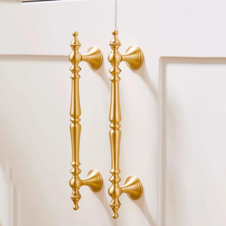 Peut inclure: Deux poign&eacute;es de placard dor&eacute;es avec un design orn&eacute;. Chaque poign&eacute;e a une longue poign&eacute;e &eacute;lanc&eacute;e avec des d&eacute;tails d&eacute;coratifs et une base arrondie. Les poign&eacute;es sont mont&eacute;es sur une porte d'armoire blanche.