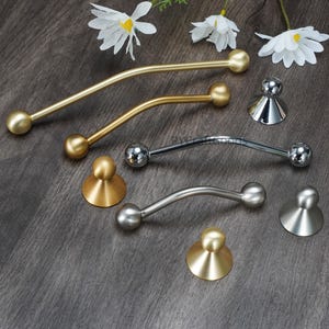 May include: An assortment of cabinet hardware, including curved bar pulls and knob pulls. The pulls are in gold and silver tones, with a spherical design at each end. The knobs have a conical base and a spherical top. White flowers are in the background.
