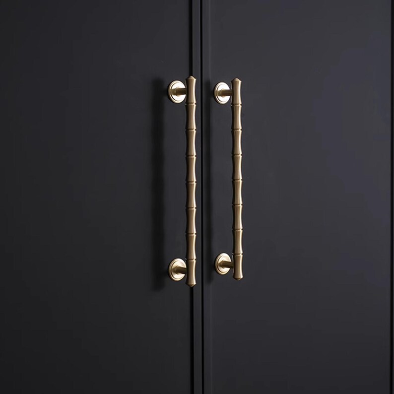 May include: Two brass-coloured cabinet pulls with a bamboo design are mounted on a dark grey cabinet door. The pulls are vertically oriented, with a circular base and a long, textured handle. The bamboo-style handles add a touch of nature-inspired elegance.