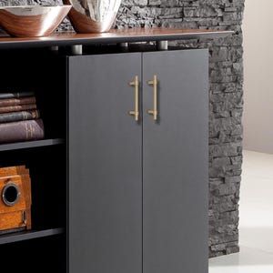 K&ouml;nnte beinhalten: Ein dunkelgrauer Schrank mit zwei T&uuml;ren und goldfarbenen Griffen. Der Schrank ist Teil eines gr&ouml;&szlig;eren M&ouml;belst&uuml;cks mit einer dunklen Holzplatte und einer Steinmauer im Hintergrund. Regale auf der linken Seite beherbergen B&uuml;cher und eine Vintage-Kamera.