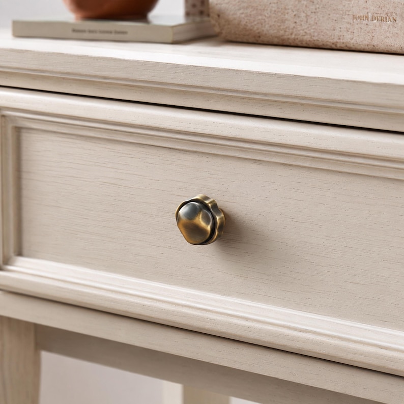 May include: Close-up of a light-coloured wooden drawer with a decorative, antique-style brass knob. The drawer is part of a larger piece of furniture, possibly a bedside table or dresser, with visible wood grain.