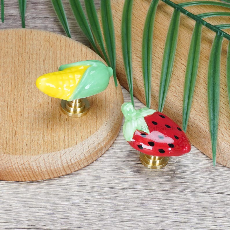 Pode incluir: Dois puxadores decorativos para arm&aacute;rios. Um &eacute; um design de espiga de milho amarelo e verde. O outro &eacute; um morango vermelho com sementes pretas e um caule verde. Ambos os puxadores t&ecirc;m uma base de lat&atilde;o.