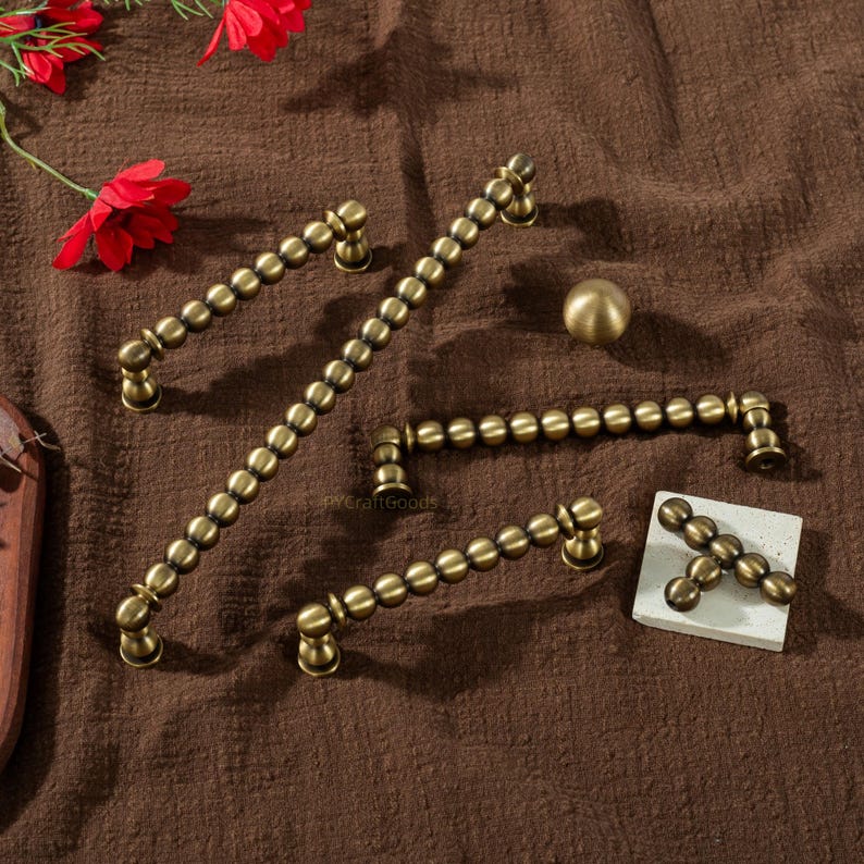 K&ouml;nnte beinhalten: Eine Auswahl an antiken Messing-Schrankbeschl&auml;gen, darunter Griffe und ein Knauf, auf einer braunen Oberfl&auml;che angeordnet. Die Beschl&auml;ge weisen ein perlenbesetztes Design auf und sind f&uuml;r M&ouml;bel oder Schr&auml;nke geeignet.