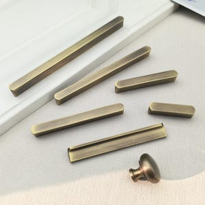 May include: An assortment of antique brass cabinet hardware, including long bar pulls, short pulls, and a round knob. The hardware has a warm, aged finish, and is arranged on a light-colored surface.