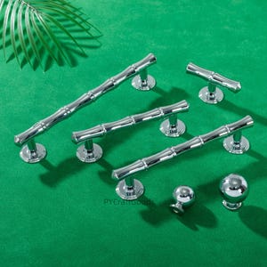 May include: A selection of polished chrome cabinet hardware, featuring bamboo-style handles and spherical knobs. The hardware is presented on a green surface, with a palm leaf in the background. The handles are of varying lengths.