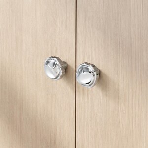 May include: Two polished chrome cabinet knobs with a distinctive, multi-faceted design. They are attached to a light-toned wooden cabinet door, presenting a contemporary and stylish appearance. These knobs are a decorative hardware feature.