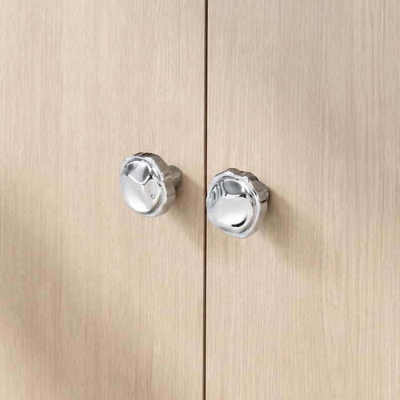 May include: Two polished chrome cabinet knobs with a distinctive, multi-faceted design. They are attached to a light-toned wooden cabinet door, presenting a contemporary and stylish appearance. These knobs are a decorative hardware feature.