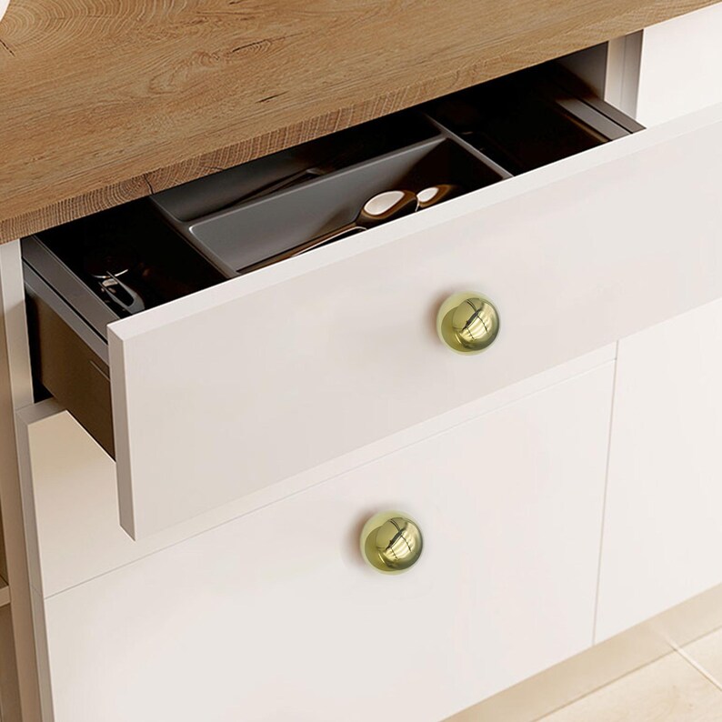 K&ouml;nnte beinhalten: Wei&szlig;er K&uuml;chenschrank mit einer ge&ouml;ffneten Schublade, die Besteck in einem grauen Organizer zeigt. Der Schrank hat eine helle Holz-Arbeitsplatte und runde, goldfarbene Kn&ouml;pfe.