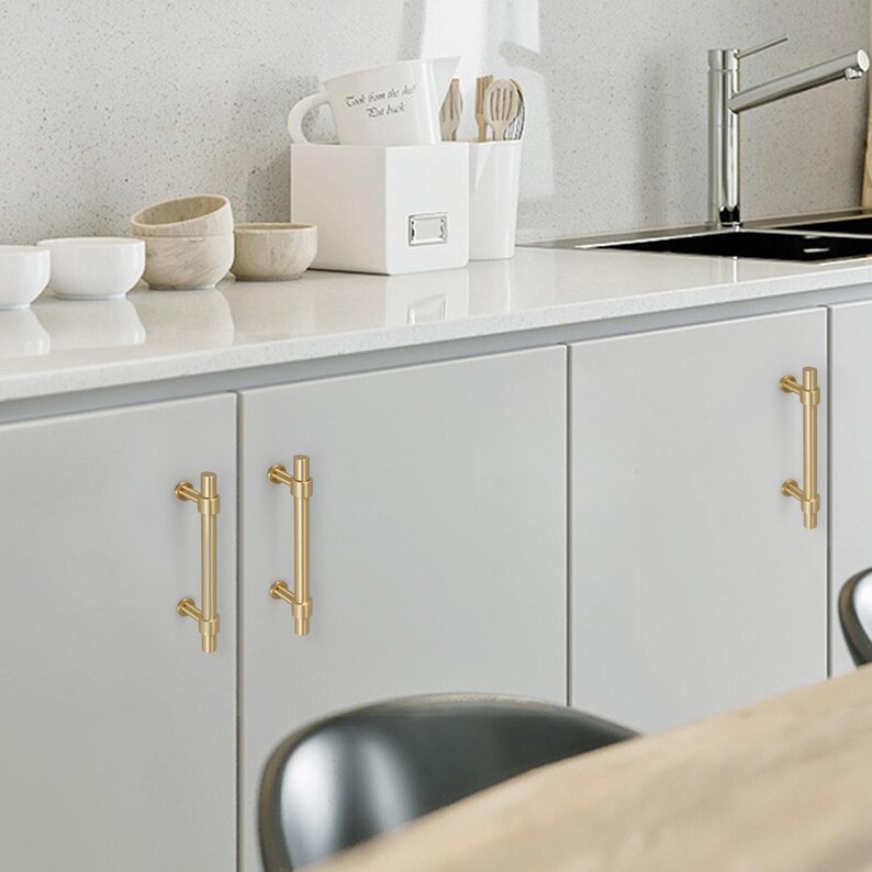 K&ouml;nnte beinhalten: Helle, graue K&uuml;chenschr&auml;nke mit goldfarbenen Stangengriffen. Die Arbeitsplatte ist wei&szlig;, und es gibt wei&szlig;e Schalen und einen wei&szlig;en Beh&auml;lter mit Utensilien auf der Theke. Die Griffe sind etwa 15 cm lang.