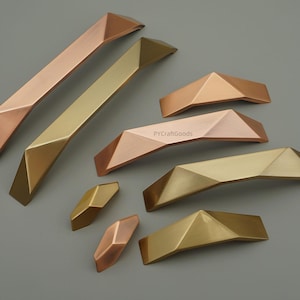 May include: An assortment of geometric cabinet pulls in various metallic finishes, including copper, gold, and brass. The pulls have a faceted design, with angular planes creating a modern aesthetic. The hardware is arranged on a gray background.