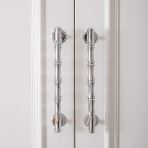 May include: Two polished chrome cabinet pulls with a bamboo-inspired design. The pulls are vertically oriented and mounted on a white cabinet door. The bamboo design adds a touch of nature-inspired elegance to the hardware.