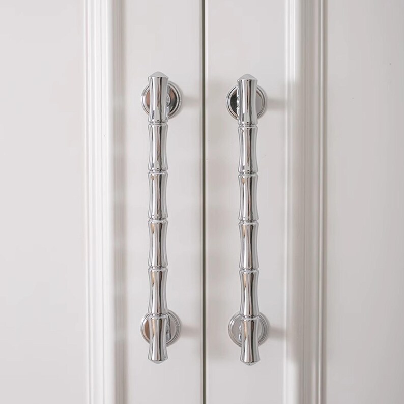 May include: Two polished chrome cabinet pulls with a bamboo-inspired design. The pulls are vertically oriented and mounted on a white cabinet door. The bamboo design adds a touch of nature-inspired elegance to the hardware.