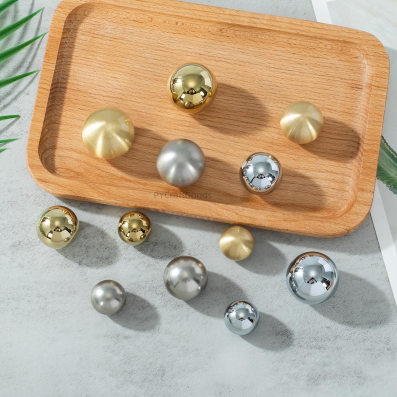 K&ouml;nnte beinhalten: Eine Sammlung dekorativer Schrankkn&ouml;pfe in verschiedenen Metallausf&uuml;hrungen, darunter Gold, Silber und Chrom. Die Kn&ouml;pfe sind kugelf&ouml;rmig und auf einem Holztablett angeordnet, das eine Reihe von Gr&ouml;&szlig;en und Stilen bietet.