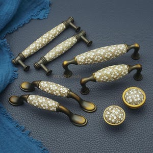 May include: Decorative cabinet hardware set featuring handles and knobs. The handles have a bronze finish with a ceramic center adorned with a white floral pattern on a tan background. The knobs have a matching design.