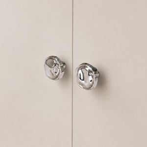 May include: Two polished silver cabinet knobs with an unusual, organic shape. The knobs are mounted on a light beige cabinet door, creating a modern and minimalist aesthetic. The knobs have a reflective surface.