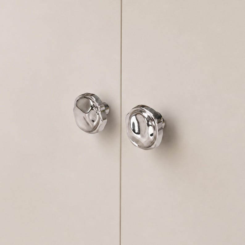 May include: Two polished silver cabinet knobs with an unusual, organic shape. The knobs are mounted on a light beige cabinet door, creating a modern and minimalist aesthetic. The knobs have a reflective surface.