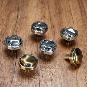 May include: Six decorative cabinet knobs with a unique, textured design. Five are silver-toned, and one is bronze-toned. The knobs are arranged on a wooden surface, showcasing their reflective surfaces and detailed craftsmanship. These knobs are ideal for furniture upgrades.