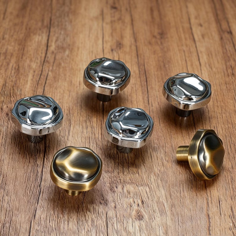 May include: Six decorative cabinet knobs with a unique, textured design. Five are silver-toned, and one is bronze-toned. The knobs are arranged on a wooden surface, showcasing their reflective surfaces and detailed craftsmanship. These knobs are ideal for furniture upgrades.